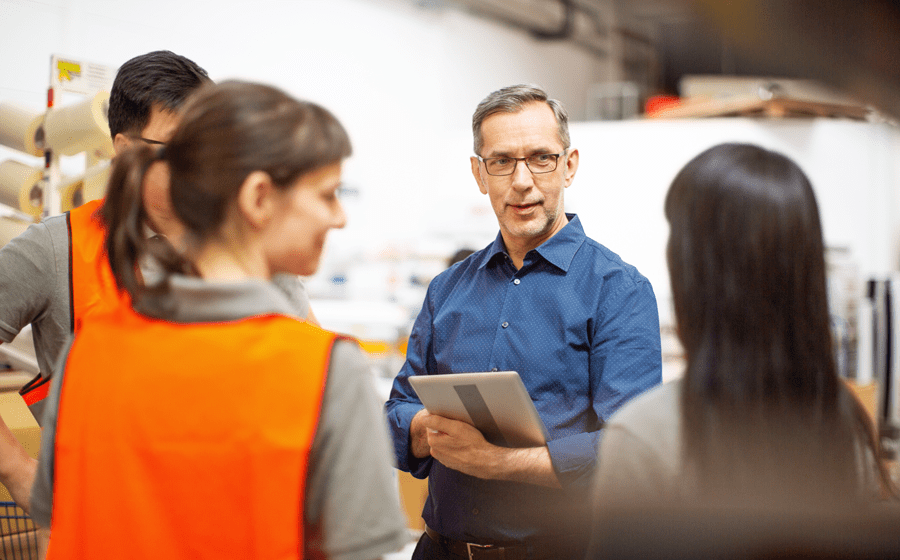 Male in blue shirt and iPad talking to one male and one female in orange vests and one female in a blue shirt in a production facility