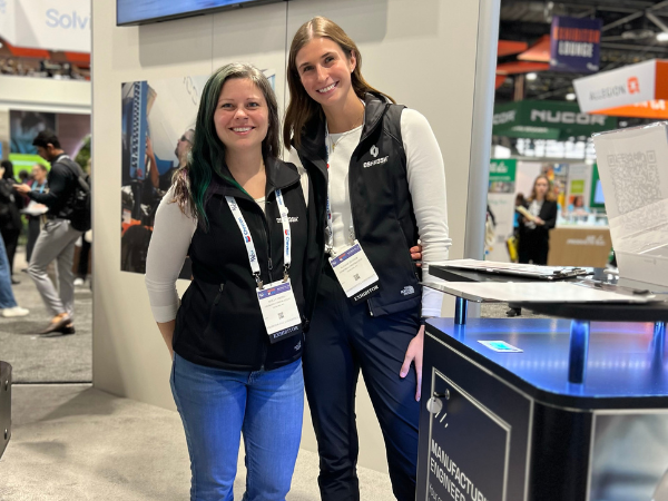 Two young females standing inside a tradeshow booth wearing black vests and jeans