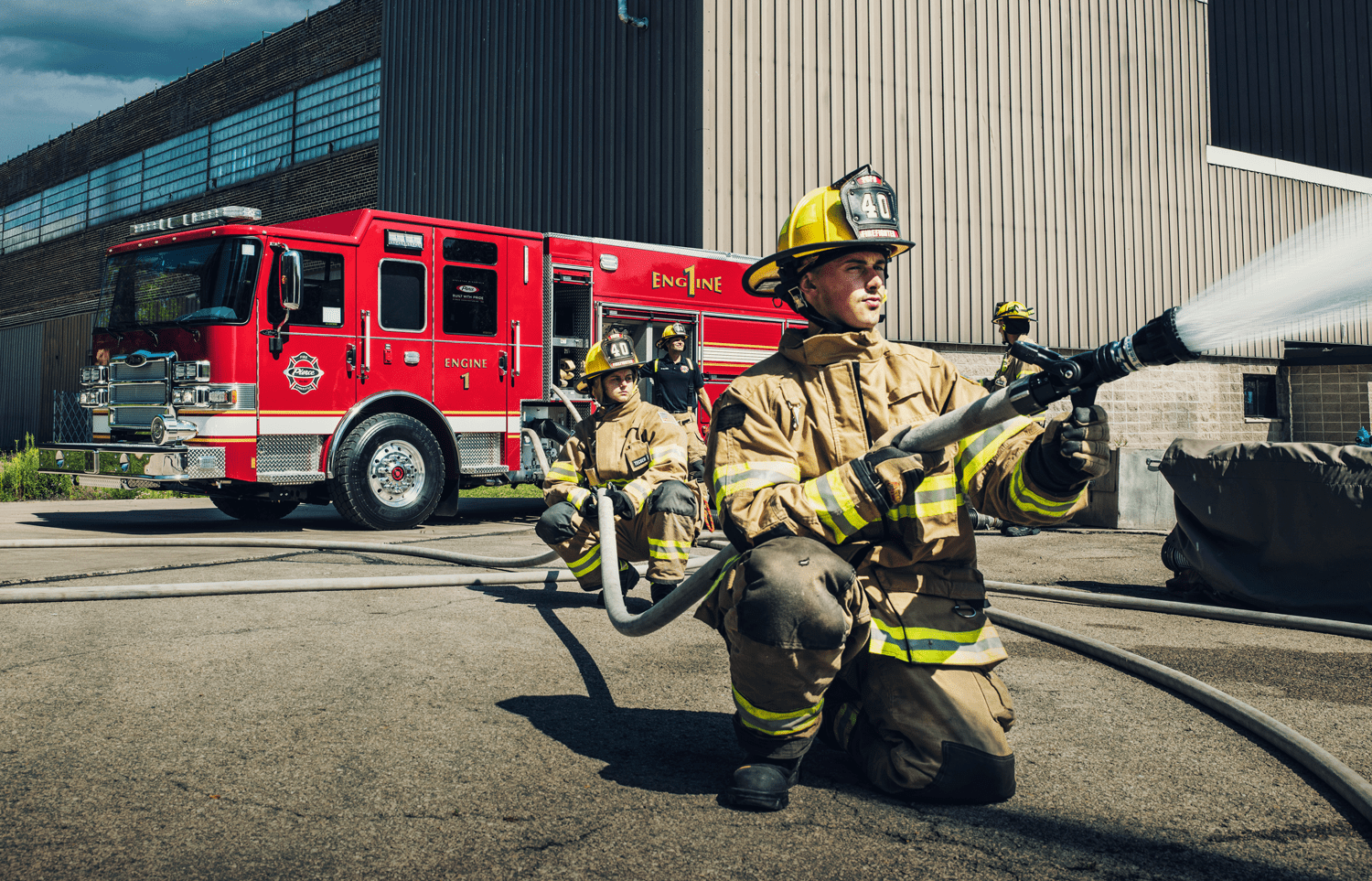 Firefighters holding a hose spraying water in front of a Red Pierce firetruck and grey building