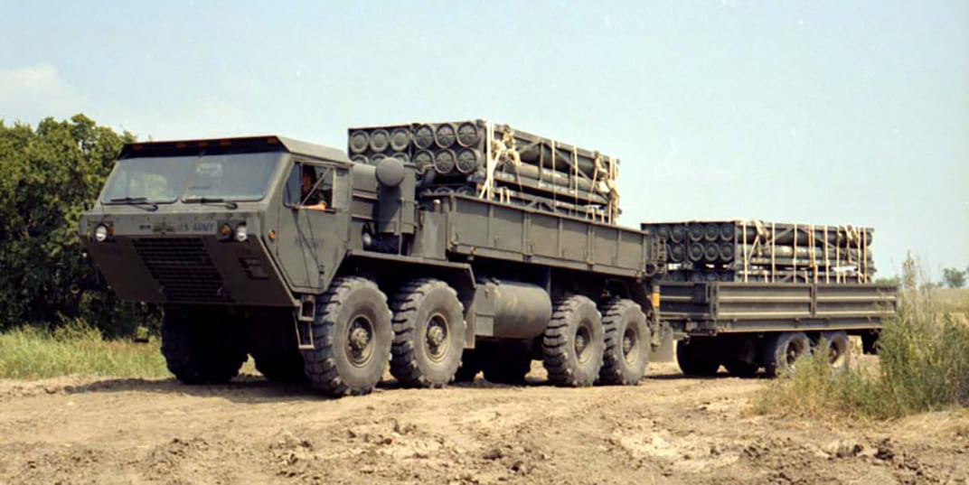 Green HEMTT carrying cargo and driving on a dirt road
