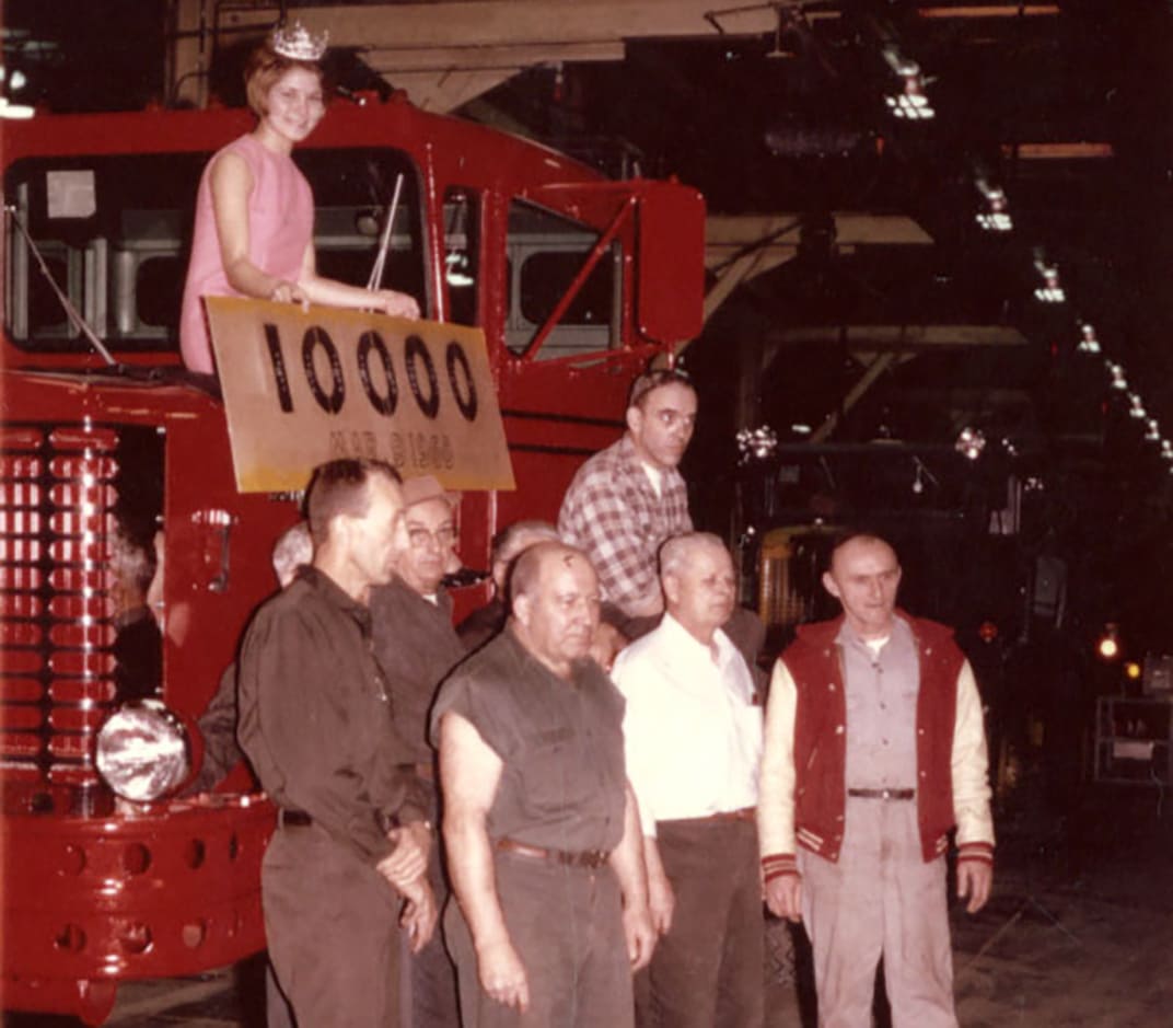 Small group of males standing in front of red 10,000th truck with a female sitting on the truck
