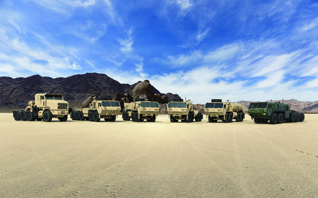Six tactical wheeled military vehicles in a row positioned outside on dirt 